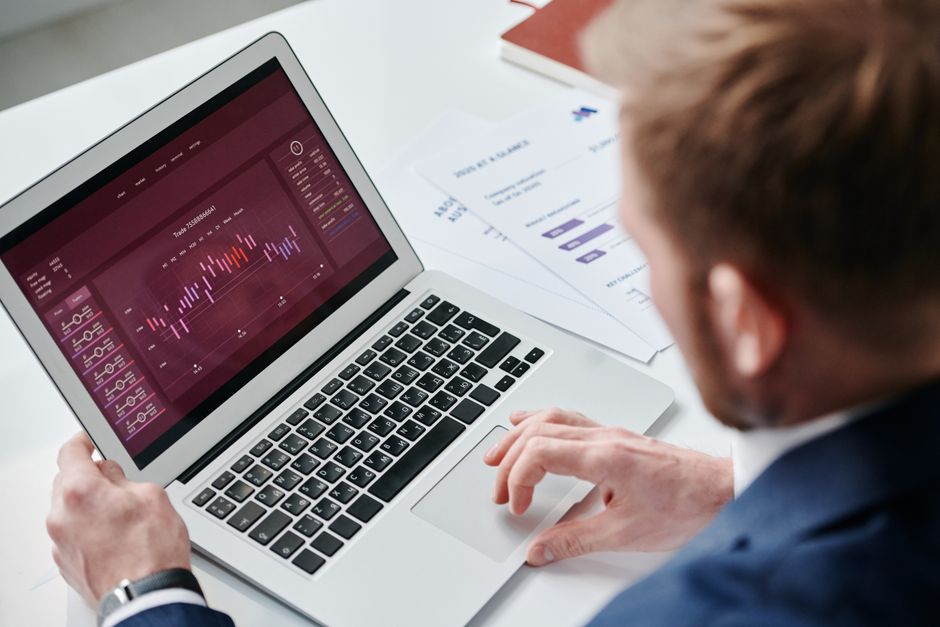Analist urmărind un tablou de bord cu grafice pe laptop, cu rapoarte tipărite alături